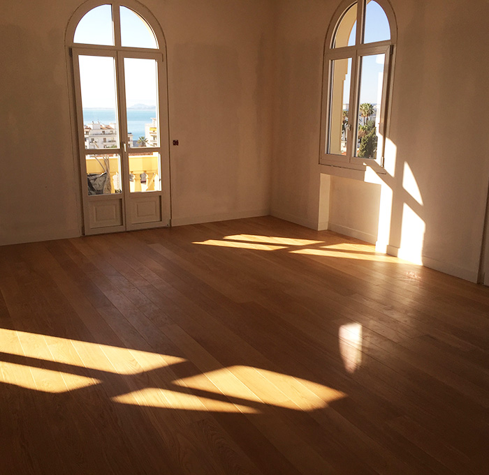 Une pièce vide avec un parquet neuf au sol, baignée de lumière du soleil qui filtre à travers les fenêtres.